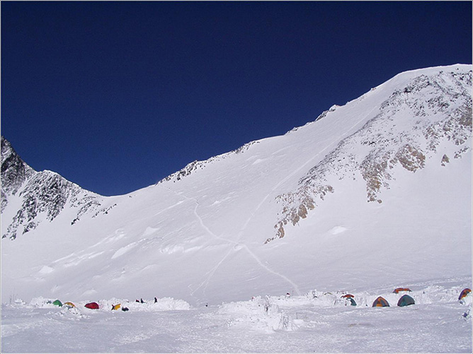 Photograph of tents in a snowfield with white mountains and a blue sky