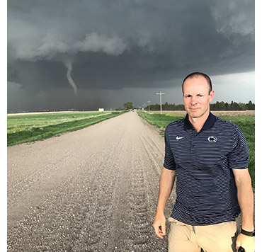 A white man in a blue polo shirt stands in front of a green field with dark gray clouds and a tornado in the distance